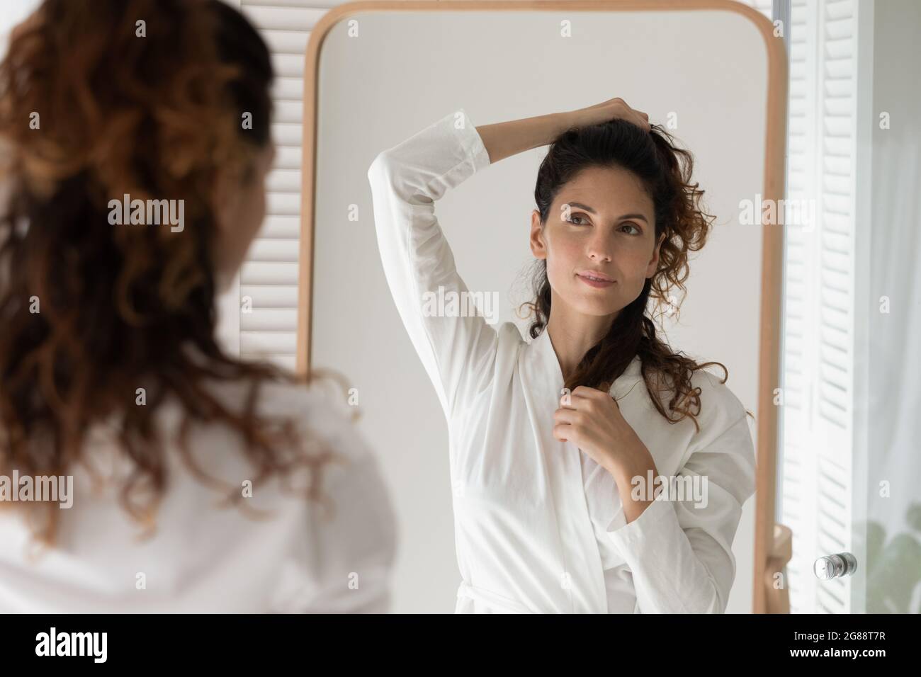 Joven mujer latina en el albornoz mirando espejo que crea el pelo Foto de stock