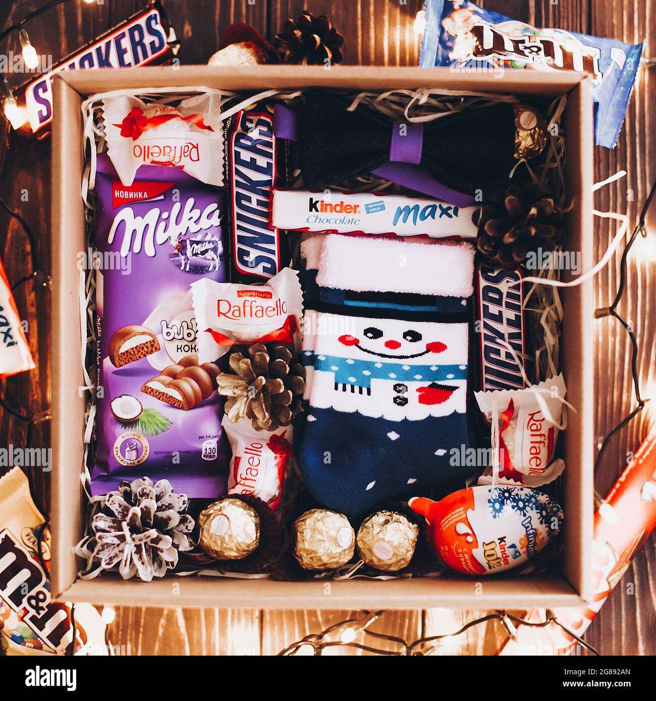 Lviv, Ucrania - 1 de diciembre de 2017: Calcetines con estilo, pajarita,  mika, snickers, kinder en paja en caja de regalo con luces de navidad.  Vista superior. Caja de regalo para c