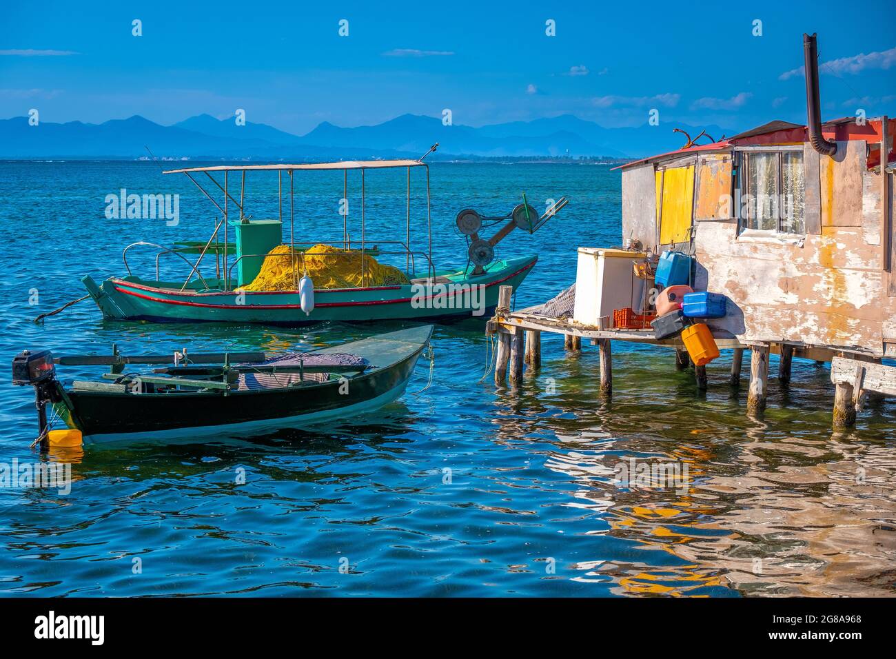 Pequeños barcos de pesca mediterráneos azules, blancos y amarillos, iluminados por el sol, con un mar turquesa brillante y un cielo azul pálido. Cabaña junto a la playa. Foto de stock