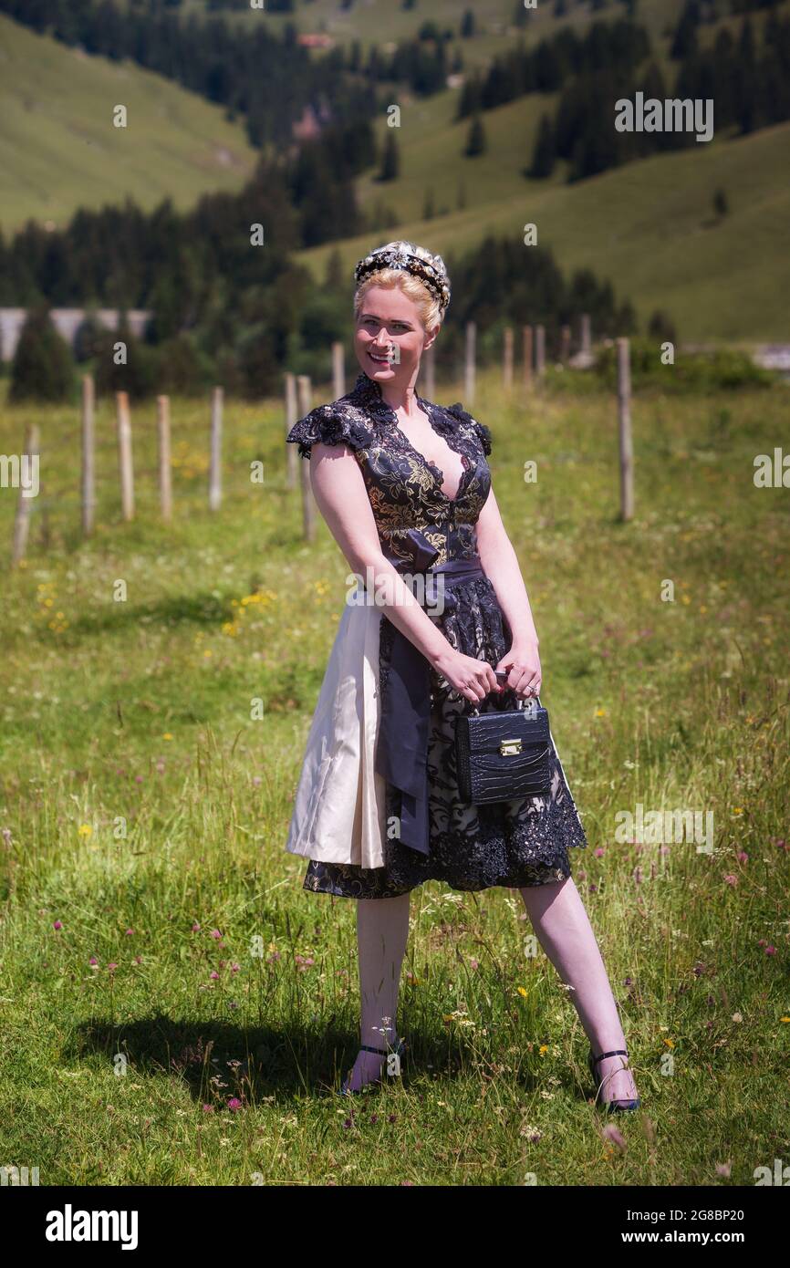 Mujer rubia en dirndl de encaje de diseñador negro con escote abierto y bolsa en un prado de montaña Foto de stock