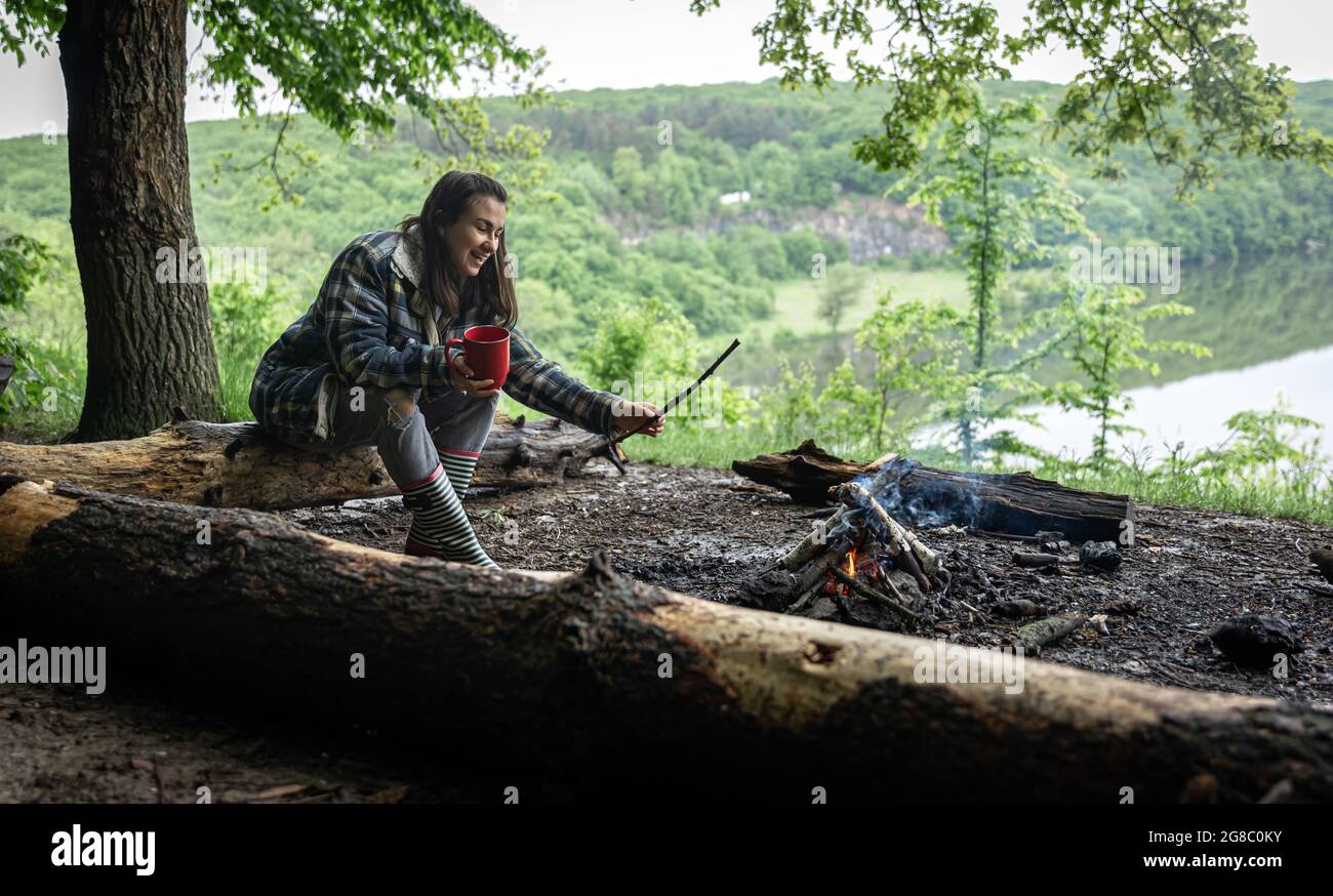 Una chica atractiva con una taza en su mano se sienta en un tronco y se calienta cerca de un fuego en el bosque. Foto de stock