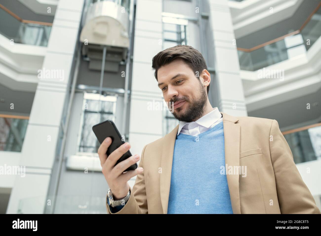 Joven hombre de negocios leyendo un mensaje en el teléfono móvil mientras se encuentra en el pasillo de la oficina Foto de stock