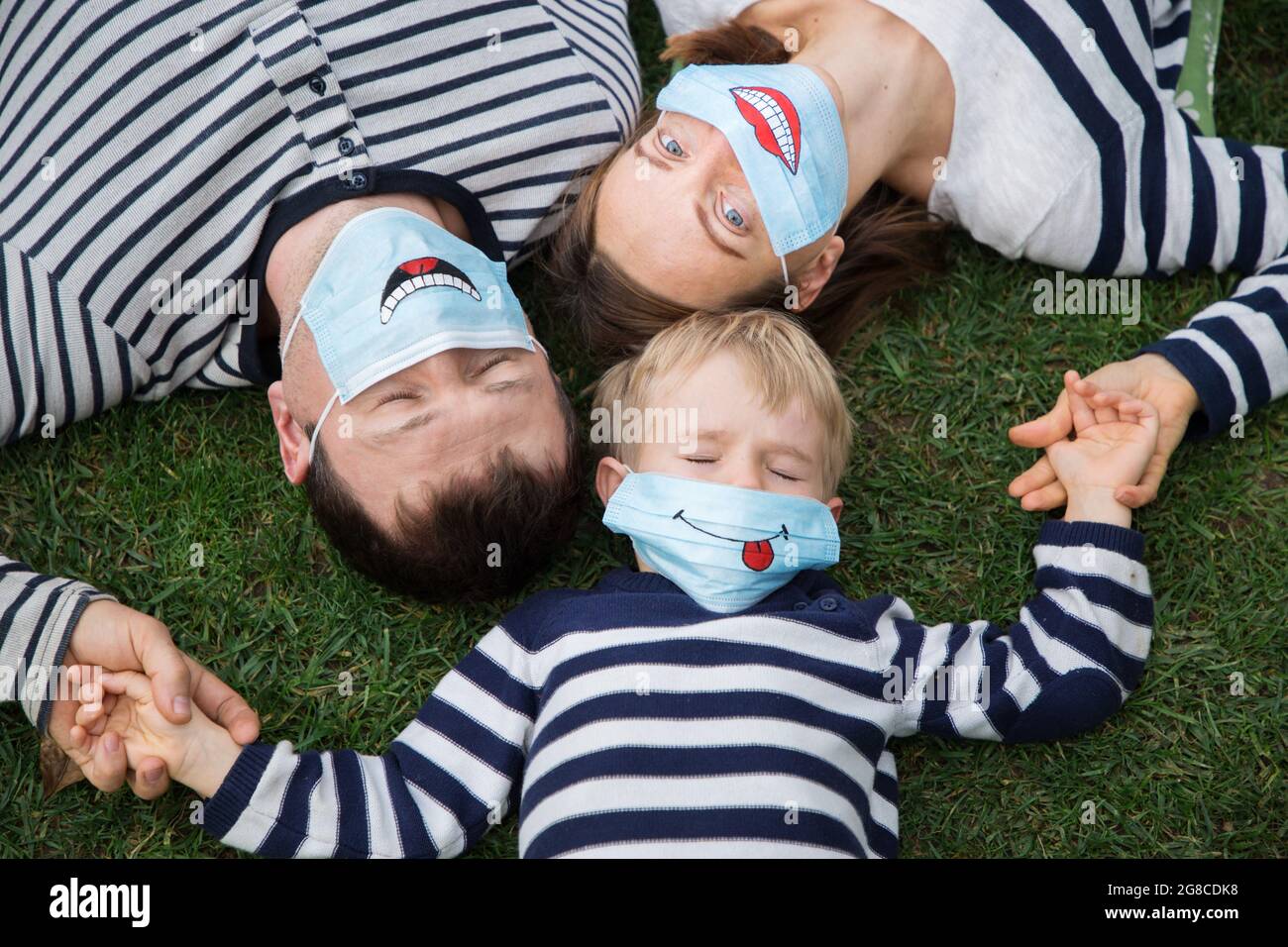 familia - hombre, mujer y niño en jerséis a rayas yacen en la hierba,  sosteniendo las manos. Están usando máscaras médicas azules con sonrisas  graciosas pintadas. Positivo Fotografía de stock - Alamy