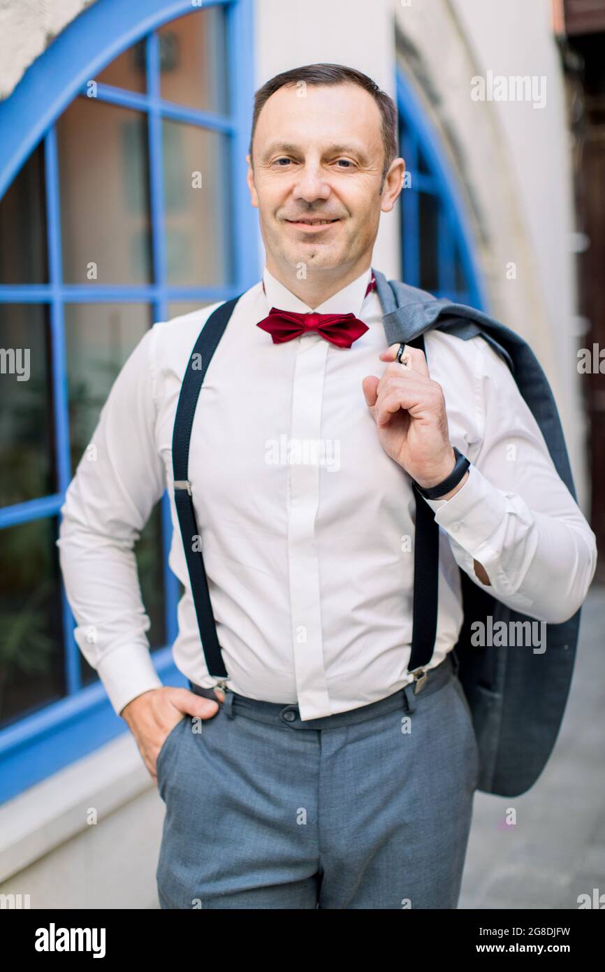 Guapo hombre caucásico maduro novio en una camisa blanca con lazo rojo y en  tirantes sostiene una chaqueta, posando en frente de un edificio vintage  con una gran ventana azul al aire