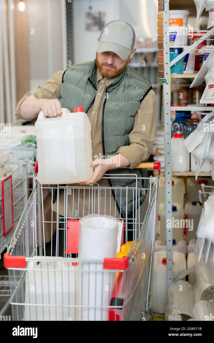 Hombre contemporáneo poniendo cisterna de plástico en el carro de la compra mientras visita la tienda de hardware Foto de stock