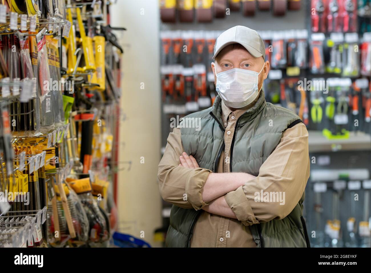 Administrador contemporáneo de la tienda de hardware en máscara protectora que cruza los brazos por el pecho Foto de stock