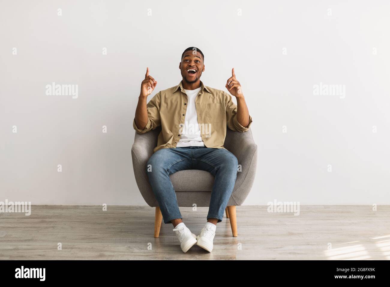Black Guy apuntando los dedos hacia arriba sentado sobre la pared gris Foto de stock