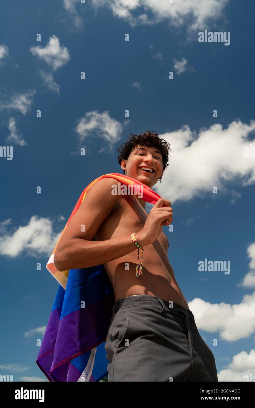 Retrato de un joven hispano gay mirando la cámara, sosteniendo la bandera LGBT - Enfoque en la cara imagen vertical Foto de stock