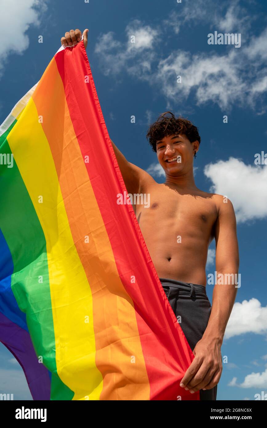 Retrato de un joven hispano gay mirando la cámara, sosteniendo la bandera LGBT - Enfoque en la cara imagen vertical Foto de stock