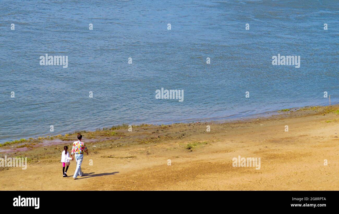 El padre y la hija caminan juntos a lo largo de la orilla del mar arenosa. El concepto de paternidad feliz y recreación familiar al aire libre. Foto de stock