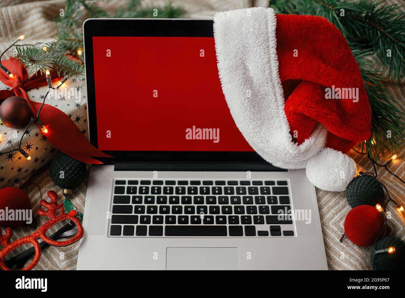 Ventas de Navidad y compras en línea. Portátil con pantalla roja vacía en sombrero de santa claus en una cómoda cama con elegantes adornos navideños, luz actual y festiva Foto de stock