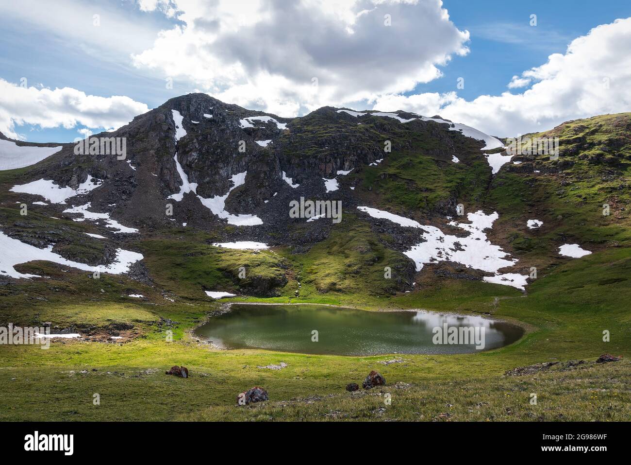 Vista increíble del lago alpino en el valle entre las montañas, nieve y hierba verde en las laderas sobre el fondo del cielo azul y las nubes. Foto de stock