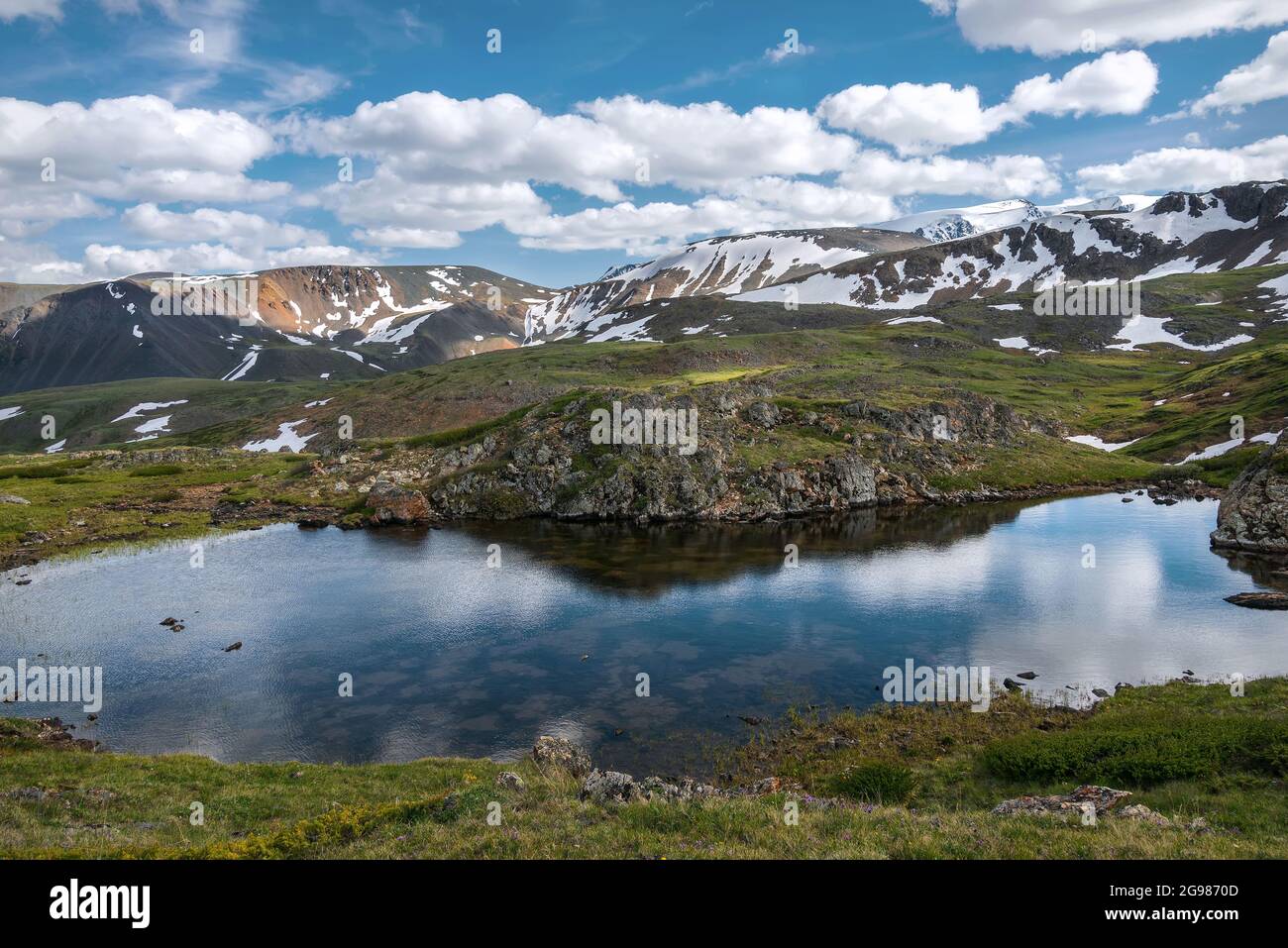 Vista increíble del lago alpino en el valle entre las montañas, nieve y hierba verde en las laderas sobre el fondo del cielo azul y las nubes. Foto de stock