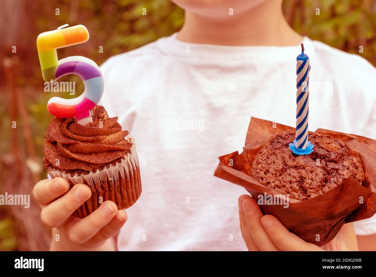 Un niño en una camiseta blanca sostiene dulces con velas en el fondo del jardín. Celebración del quinto cumpleaños. Foto de stock