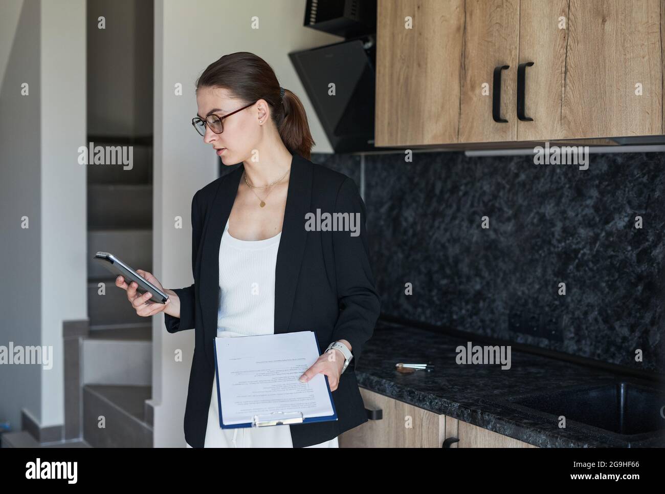 Retrato gráfico de agente inmobiliario femenino con smartphone mientras se encuentra junto a la cocina en casa para la venta, copia espacio Foto de stock