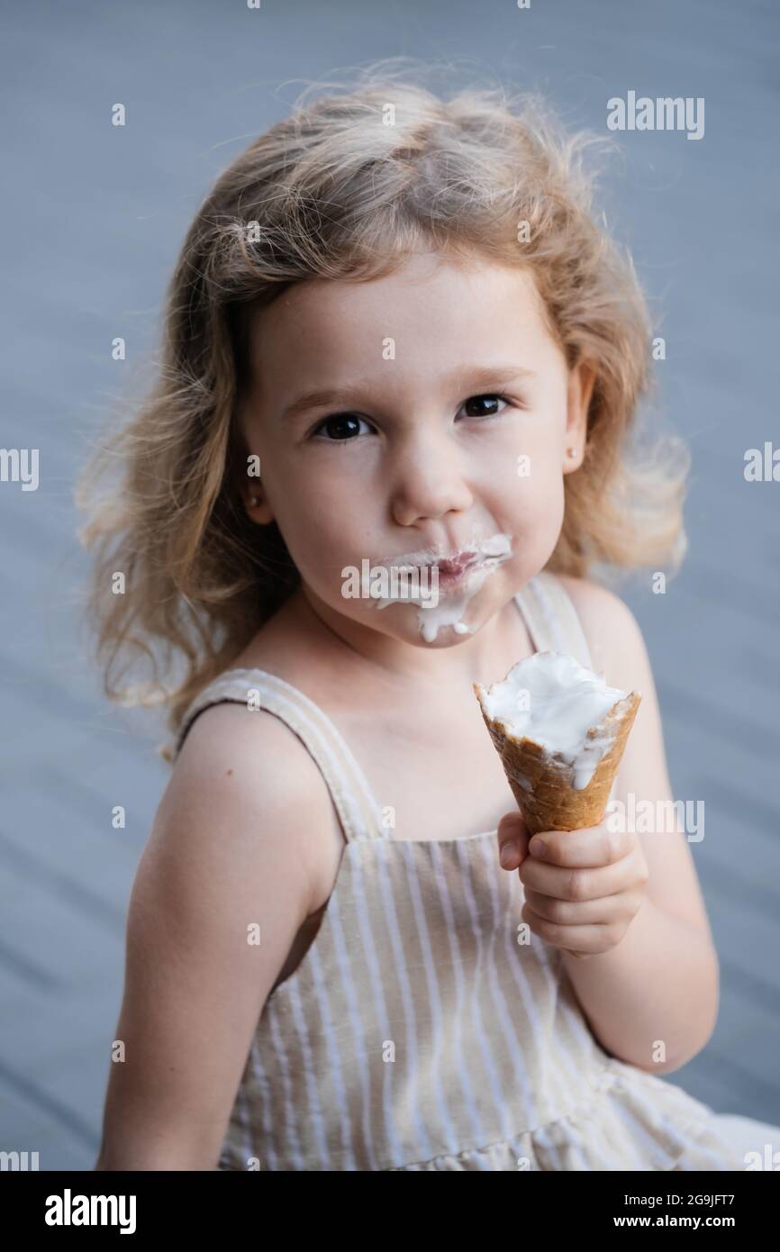 Niño pequeño comiendo helado de cono afuera cerca de la cafetería. Linda chica blanca rubia lamer comida congelada de verano en la calle. Boca sucia, niño feliz. Foto de stock