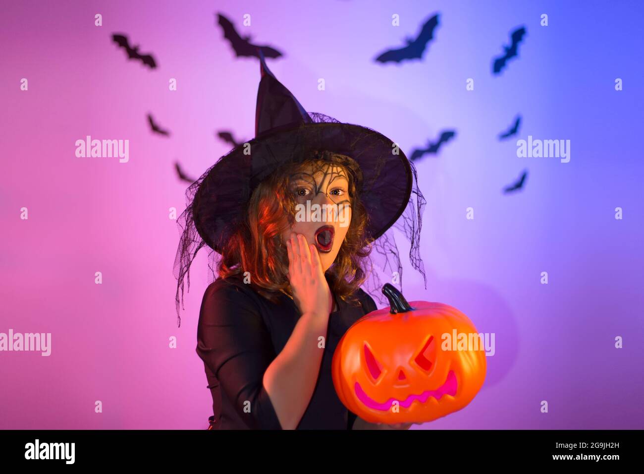 Chica divertida vestida como una bruja para Halloween con una calabaza y un  maquillaje de tela de araña en su cara. Estudio con hermosa iluminación  púrpura neón. Feliz Fotografía de stock -