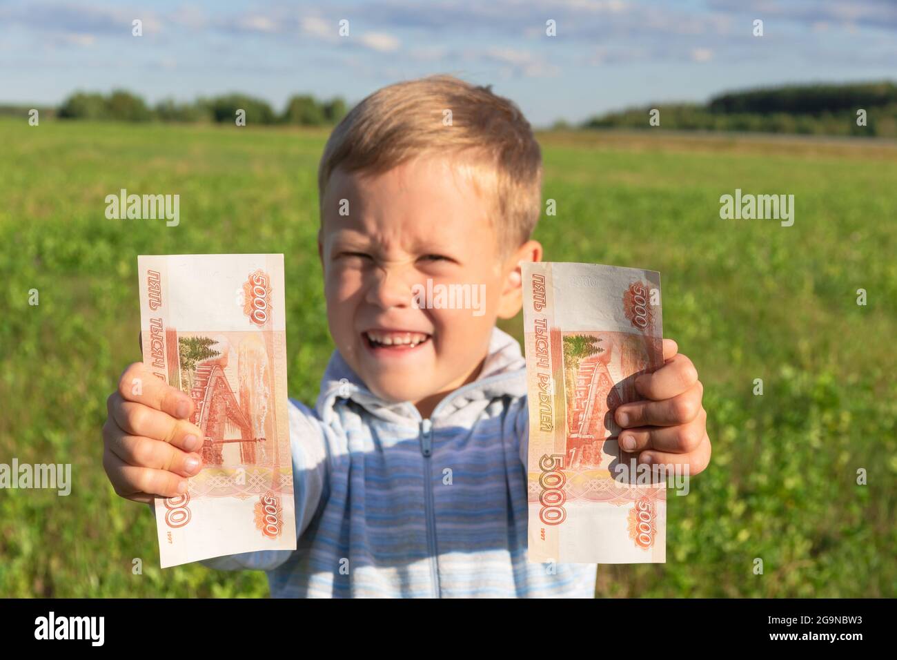 Un niño satisfecho, un niño preescolar, sostiene rublos de papel en sus manos al aire libre sobre el fondo de un campo con hierba verde en un verano soleado y brillante Foto de stock