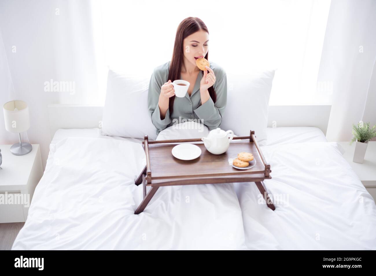 Foto de linda joven dama gris ropa de tumbada cama beber café por la mañana comer galletas sonriendo habitación Fotografía de stock - Alamy