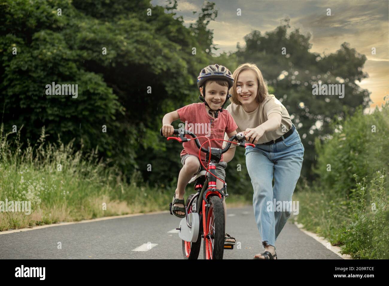 Aprender A Andar En Bici A Los 50 Madre enseñando a su hijo a montar en bicicleta. Feliz lindo niño en el  casco aprender a montar una bicicleta en el sendero de bicicletas en verano  lloviendo día a la puesta