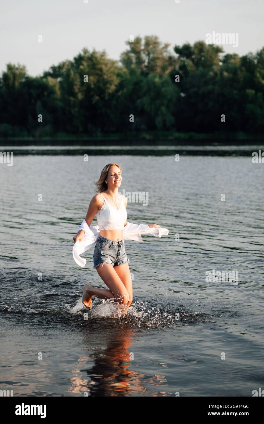 Una mujer hermosa corre libertad a través del agua. Una joven rubia  sonriente en una camiseta blanca y una camisa y pantalones cortos denim  sobre un fondo de paisaje Fotografía de stock -
