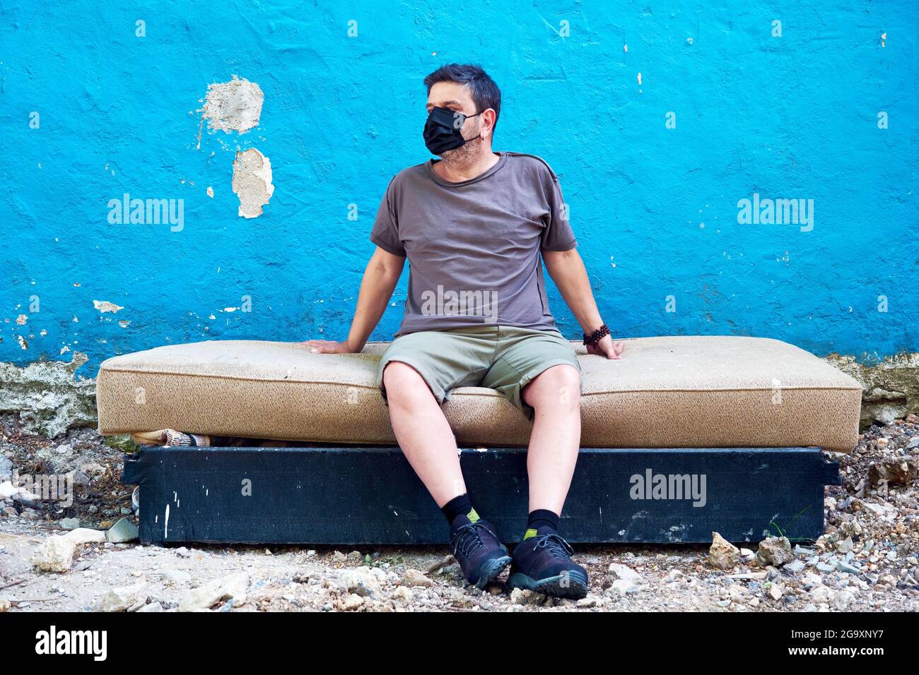 Hombre caucásico con una máscara médica protectora sentado en una cama desechada frente a una pared azul. Concepto de protección contra coronavirus y personas sin hogar. Foto de stock
