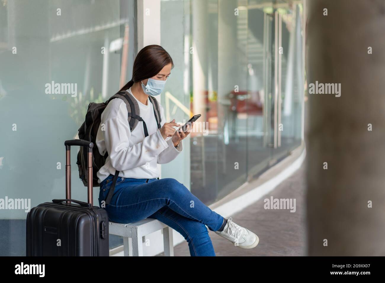 Viaje. Mujer con camisa blanca de manga larga y jeans, con una máscara.  Siéntese en la silla y utilice el teléfono móvil con la maleta colocada en  la mano derecha Fotografía de
