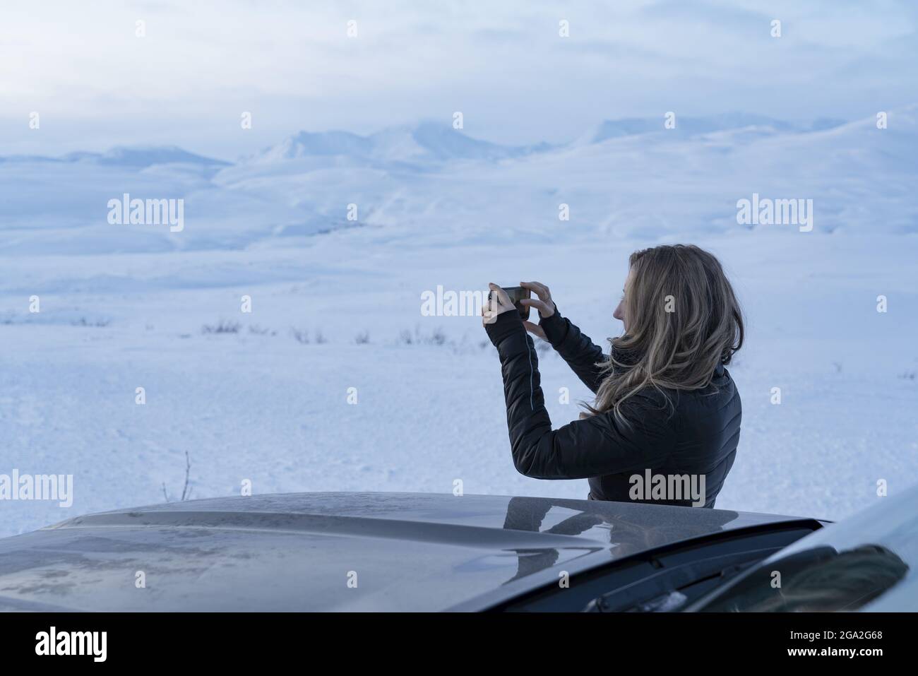 Mujer viajera de pie junto a un coche tomando una fotografía con su teléfono celular del paisaje nevado a lo largo de la autopista Haines; Yukon, Canadá Foto de stock