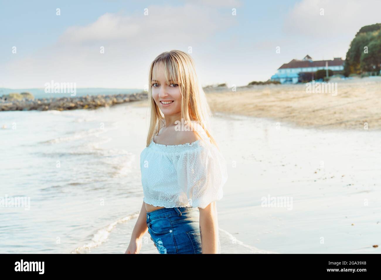 Retrato de una feliz mujer sonriente en la parte superior blanca y pantalones cortos vaqueros disfrutando del momento durante un paseo por la playa junto al mar. Vacaciones de verano, vacaciones Foto de stock