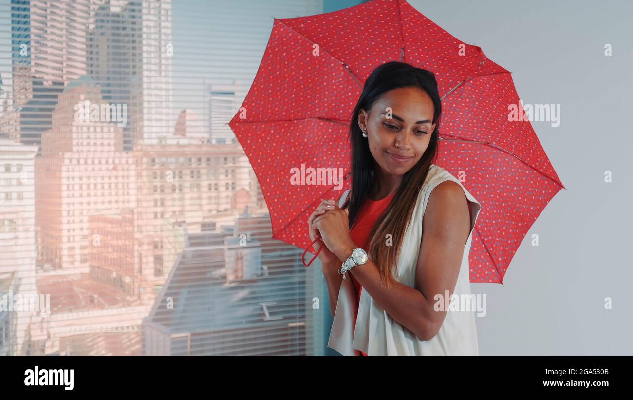 Primer plano de modelo negro sonriente posando con paraguas rojo en el bar de trona para la revista de moda de fotografía. Hay rascacielos en el fondo. Foto de stock