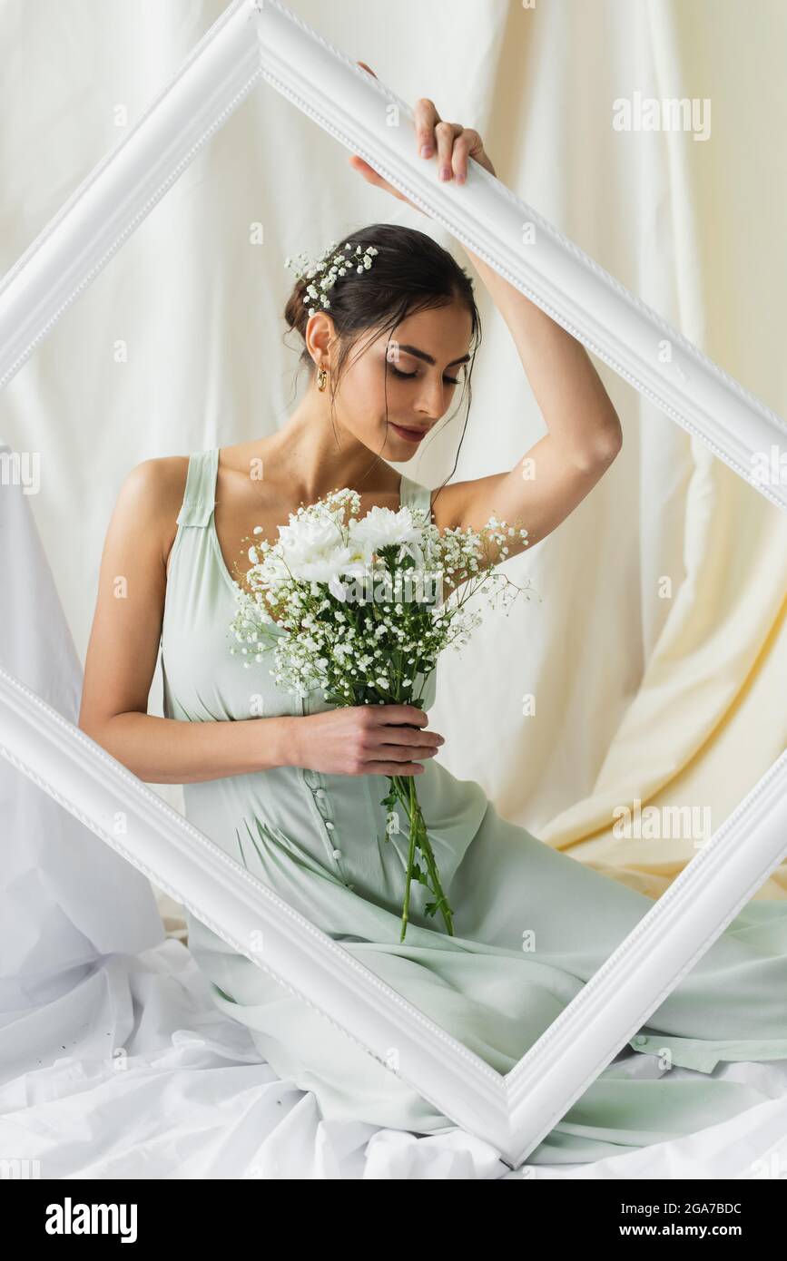 mujer sonriente sosteniendo ramo de flores florecientes y posando cerca del marco sobre blanco Foto de stock