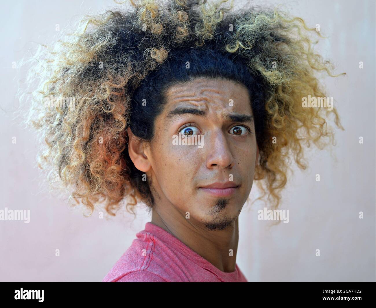 El joven latinoamericano con un choque de pelo rizado salvaje y extremos de pelo teñido rubio mira sorprendido con ojos grandes y frente arrugado en la cámara. Foto de stock
