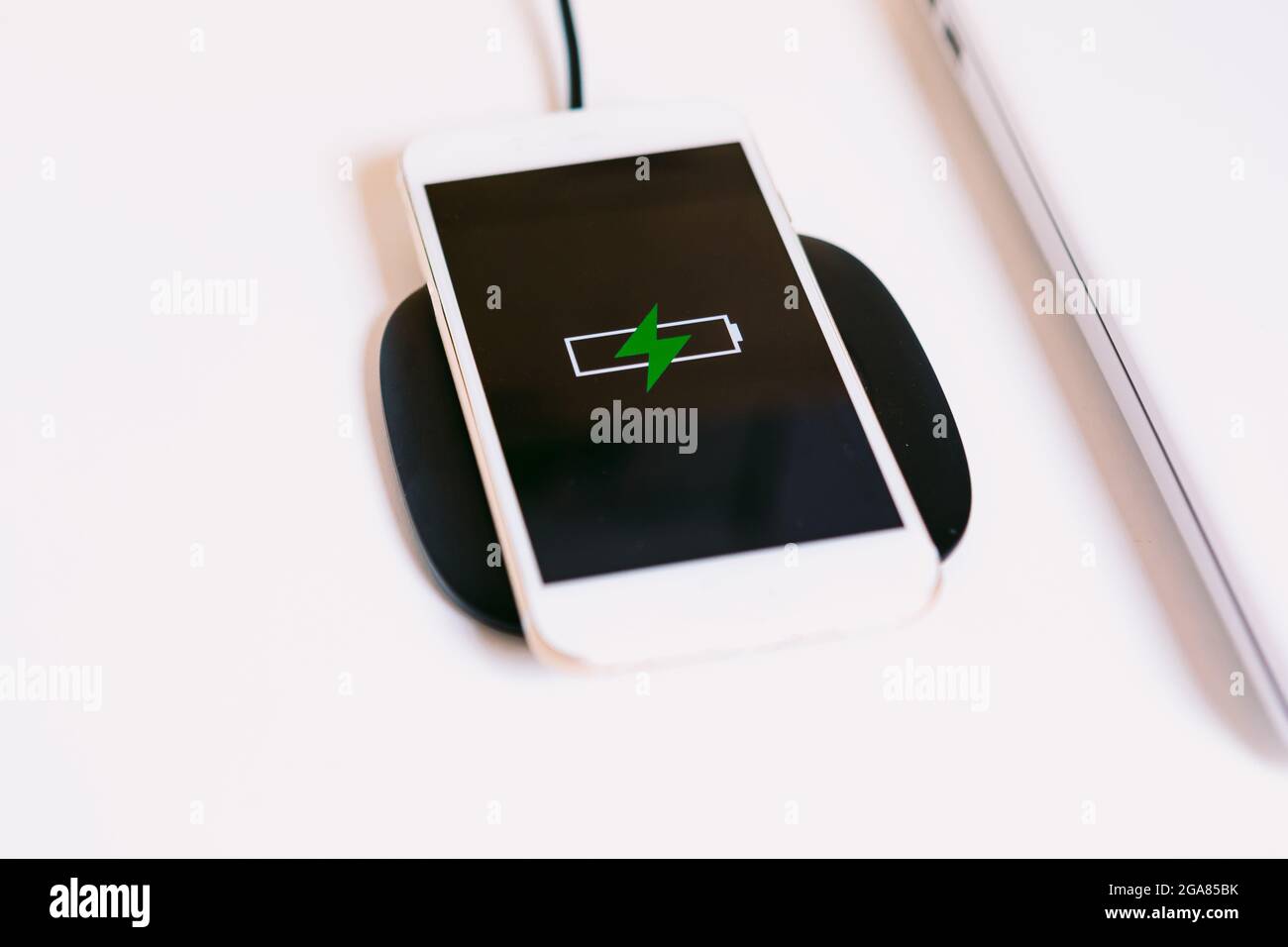 Teléfono inteligente móvil blanco con el logotipo de una batería con un  rayo verde en la pantalla, que se carga en una base de cargador sin cable  junto al portátil encendido Fotografía