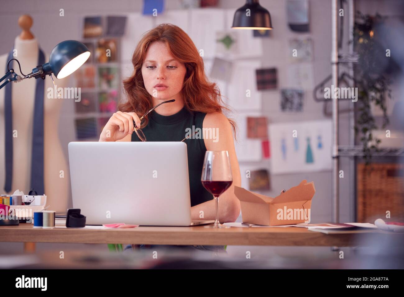 Mujer Dueño de Negocios de Moda con Copa de Vino Trabajando tarde en el portátil en el estudio Foto de stock
