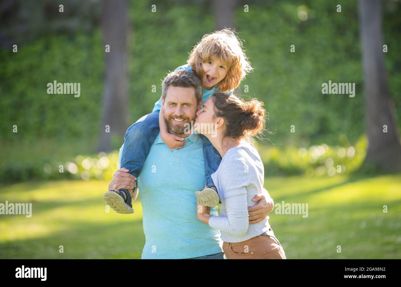 Madre beso padre mientras niño niño montar piggy detrás en los hombros de los padres verano al aire libre, familia Foto de stock