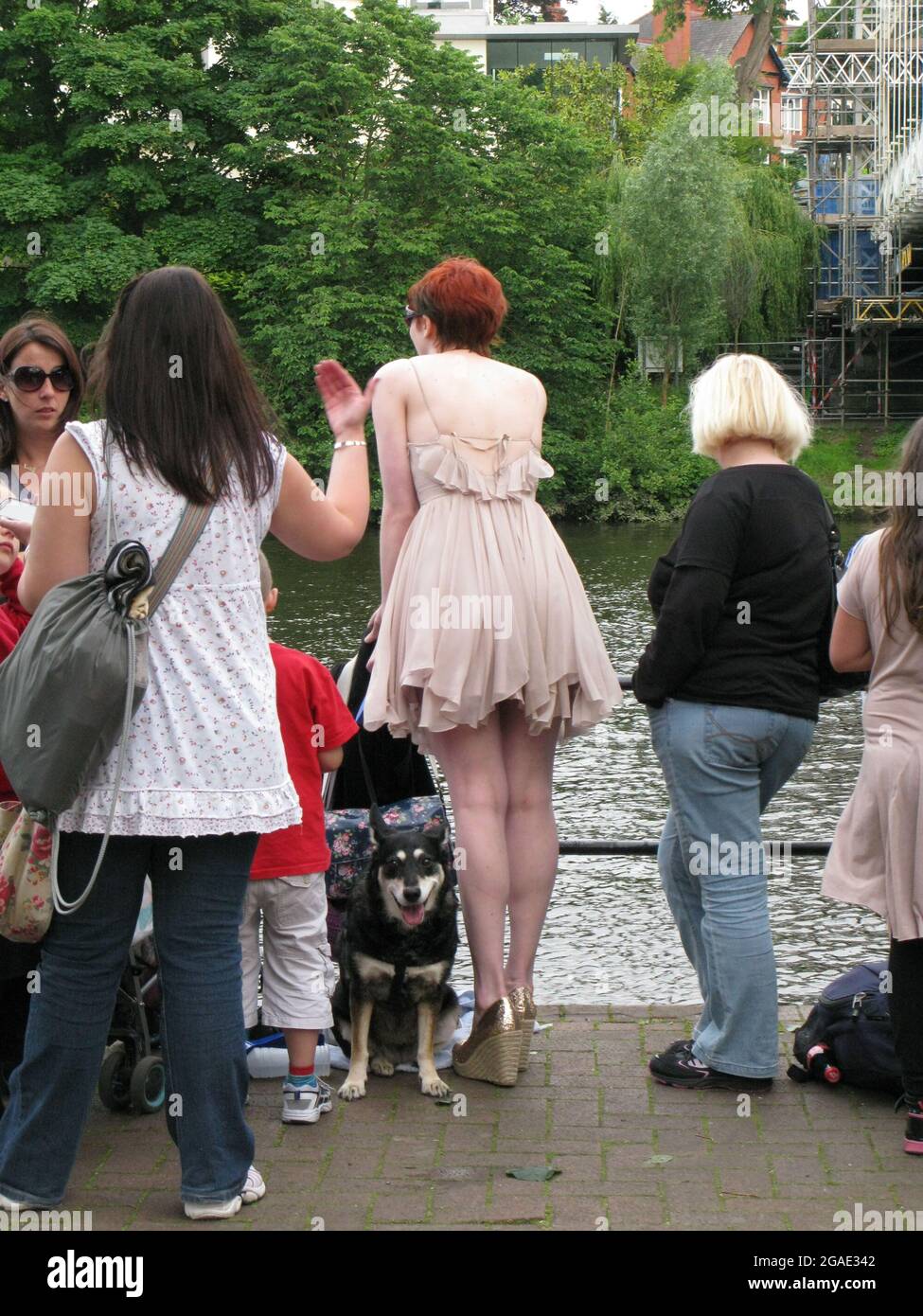 Una mujer alta y joven de cabeza roja en la orilla del río en Chester,  Cheshire, Inglaterra. Lleva un elegante vestido corto y tacones altos. Ella  tiene un perro en un plomo