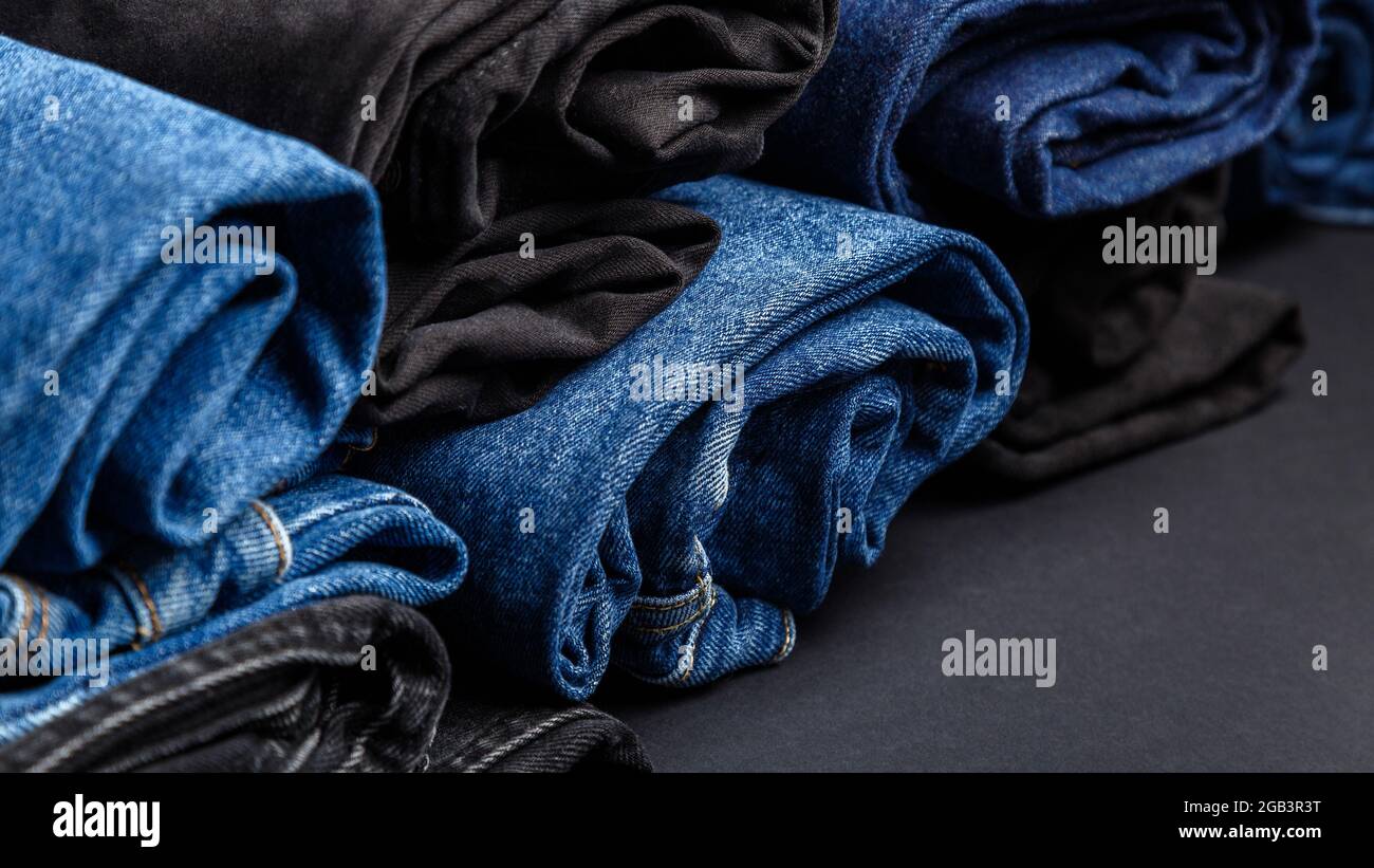 Los jeans azules doblados en rollos se encuentran sobre fondo negro.  Pantalones vaqueros pila de ropa informal. Tienda de ropa tienda Jean  textil para la venta. Banner web largo Fotografía de stock -