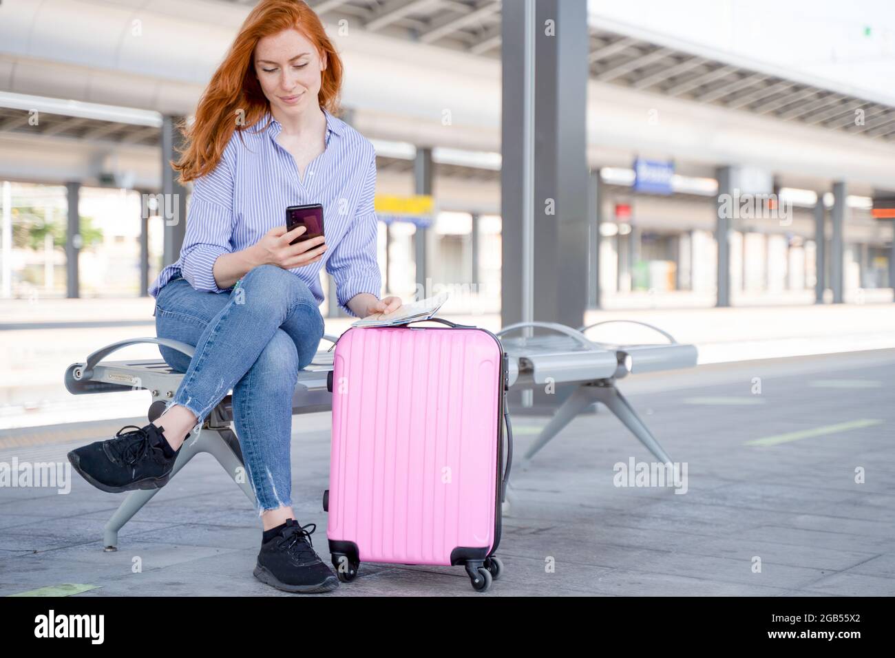 Mujer joven sosteniendo un teléfono móvil sentado en la plataforma de la estación de tren Foto de stock