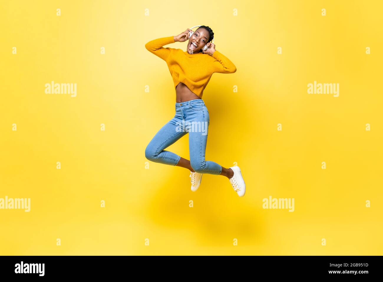 Feliz mujer joven afroamericana energética con auriculares escuchando música y saltando en un fondo de estudio aislado de color amarillo Foto de stock