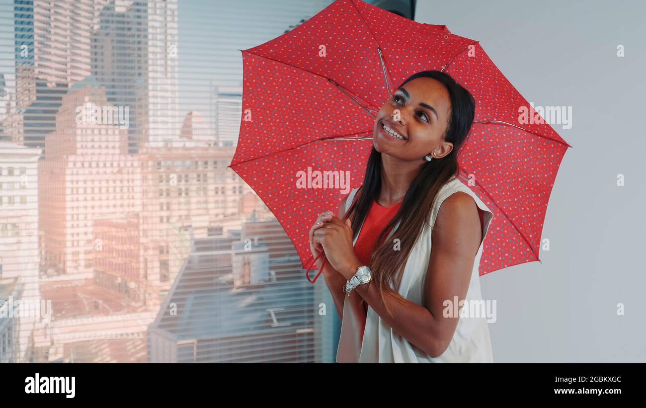 Primer plano de modelo negro sonriente posando con paraguas rojo en el bar de trona para la revista de moda de fotografía. Hay rascacielos en el fondo. Foto de stock
