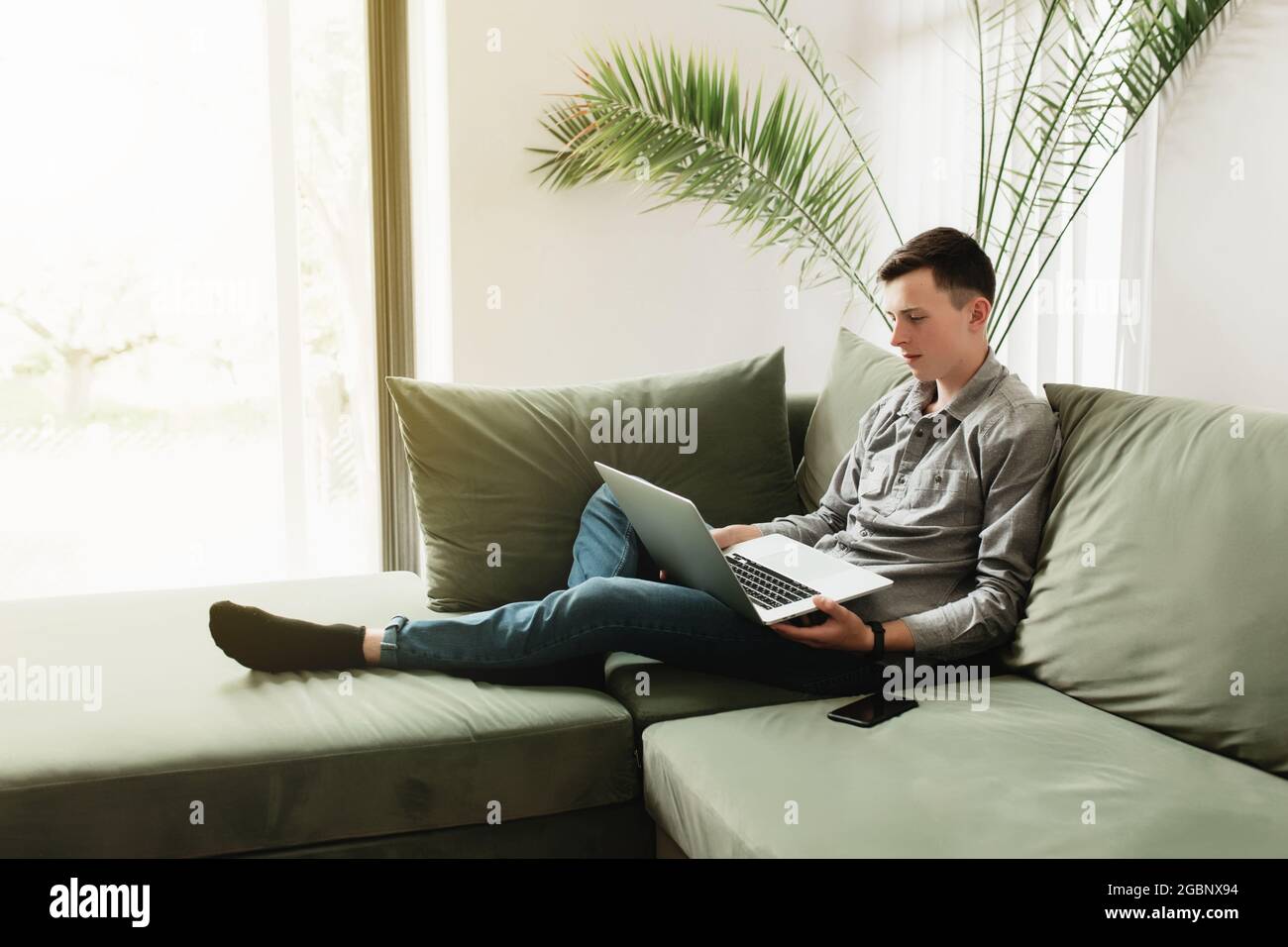 Joven sentado en un sofá verde con ordenador portátil gris, teléfono negro. El  hombre está sosteniendo un cuaderno plateado, leyendo, escribiendo algo.  Trabajo desde casa, educación CO Fotografía de stock - Alamy