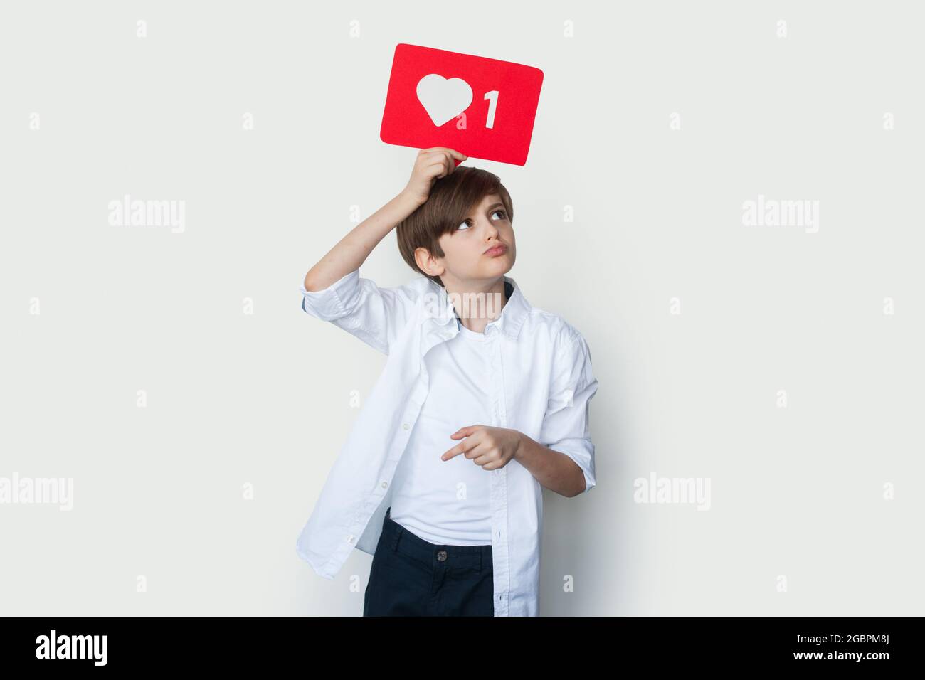 Lindo niño está sosteniendo un papel con signo similar sobre la cabeza posando en una pared blanca del estudio Foto de stock