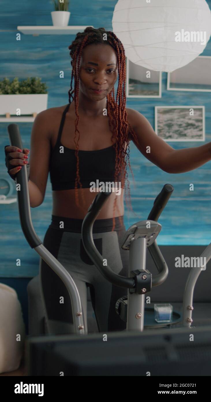 Entrenamiento atlético de la mujer negra en la bicicleta elíptica que  practica el deporte cardio que mira el vídeo aerobio en la tv durante la  rutina de la mañana del peso corporal