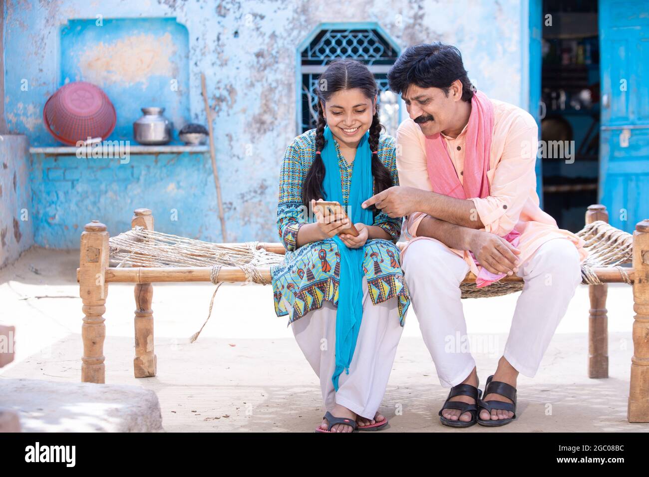 Feliz padre indio y joven hija usando smartphone mientras se sienta en la cama tradicional fuera de su casa, feliz familia rural, el hombre de la aldea que aprende Foto de stock