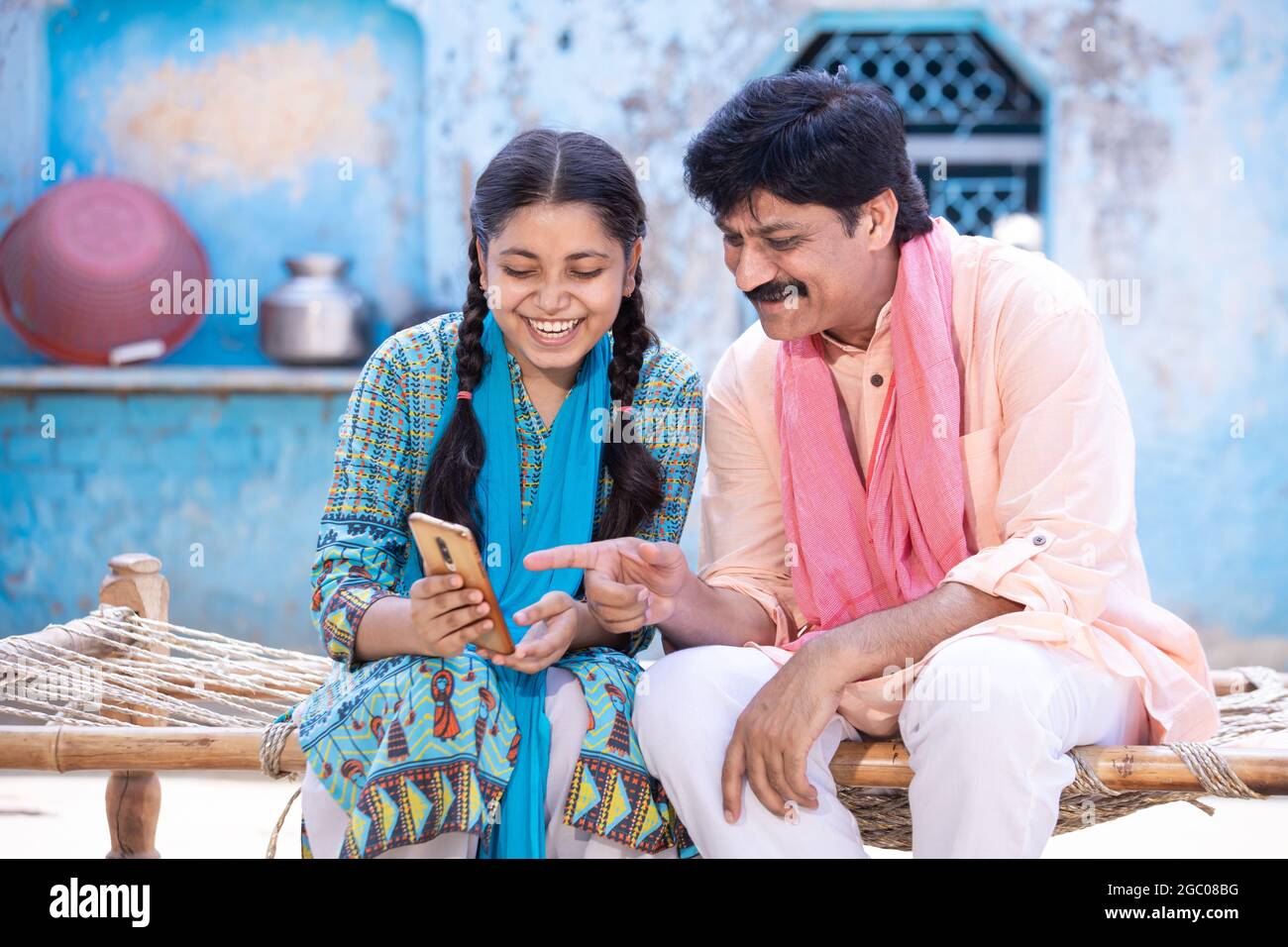 Feliz padre indio y su hija joven usando un smartphone mientras se sienta en la cama tradicional fuera de su casa, feliz familia rural, el hombre apunta con el dedo Foto de stock