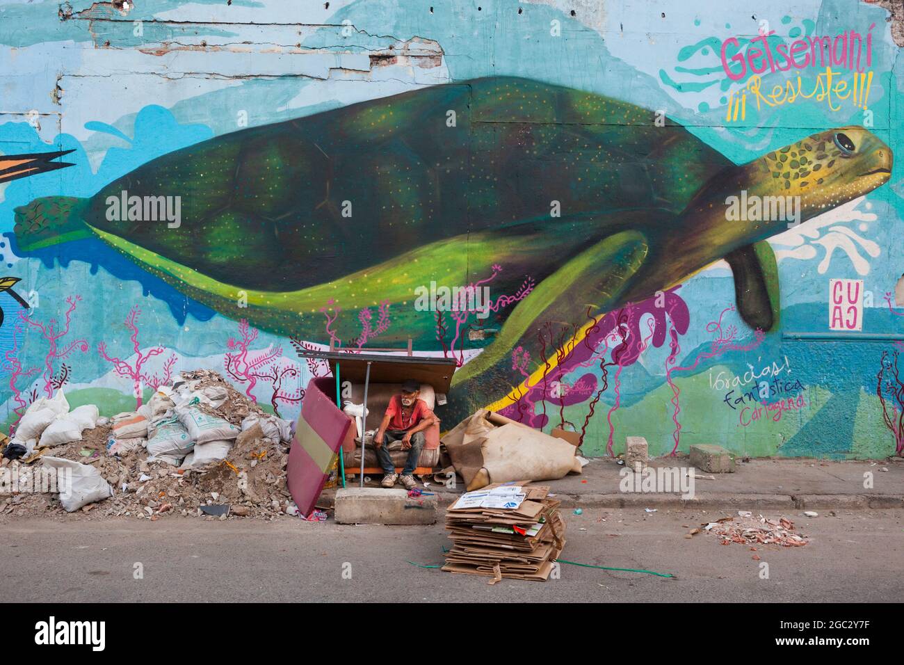 Un hombre sin hogar se sienta en un refugio de cartón improvisado junto a escombros de construcción y un mural gigante de una tortuga marina en Getsemani, Colombia. Foto de stock