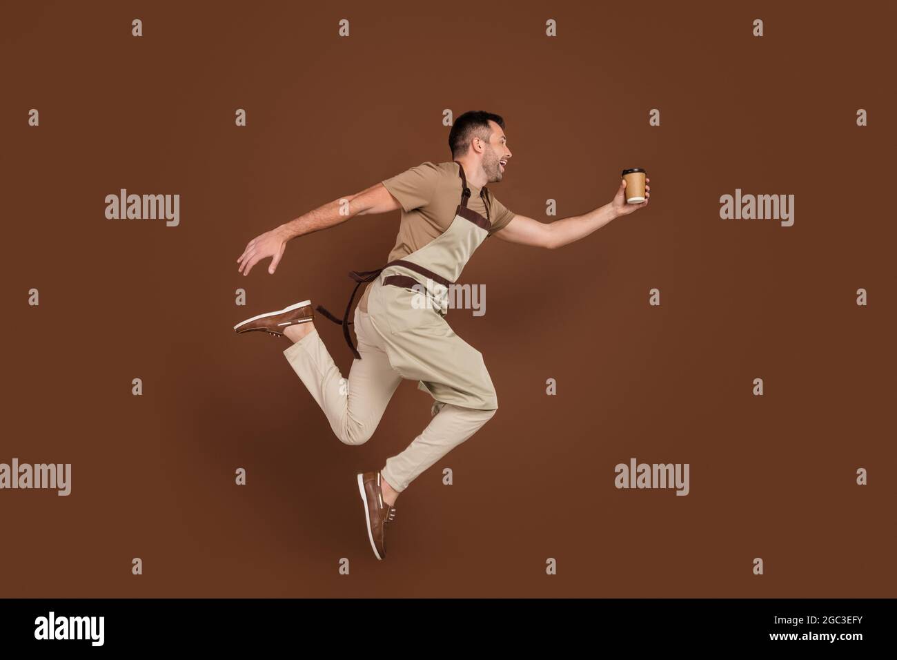 Hombre de tamaño corporal completo con delantal saltando dando una taza de café al cliente aislado color marrón de fondo Foto de stock