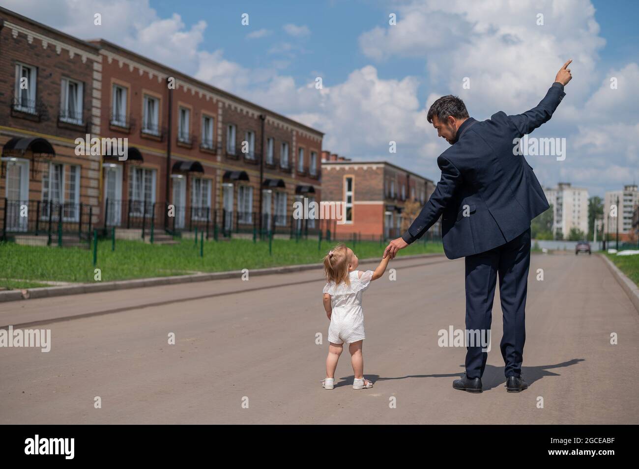 Vista trasera de un hombre en un traje negro camina con su hija pequeña al aire libre. Una niña en un vestido blanco sostiene la mano de su padre. Amor paterno Foto de stock