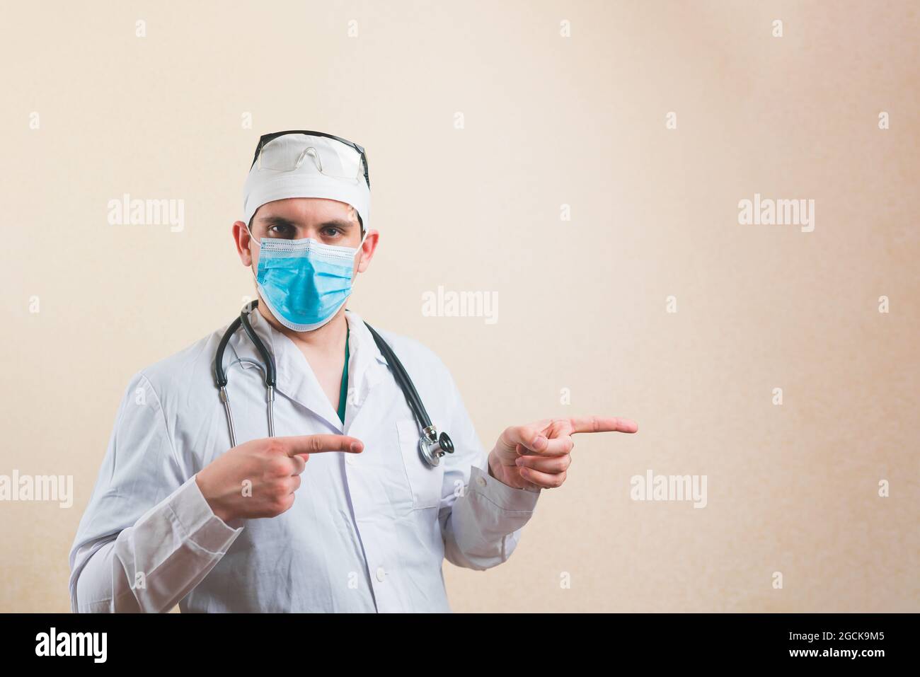 Retrato del joven doctor en traje blanco y gafas protectoras de máscara respiratoria y estetoscopio apuntando sus dedos hacia el copyspace aislado Foto de stock