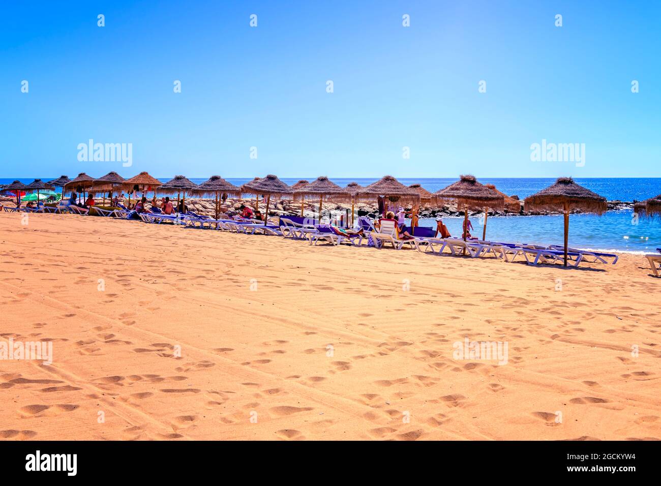 Parasoles de paja con la gente tomando el sol en las tumbonas de playa de arena en praia rosa branca Quarteira playa Quarteira Algarve Portugal Foto de stock